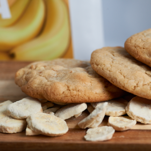 Stack of golden-brown cookies on a wooden surface with broken cookie pieces scattered around; a banana box is blurred in the background.